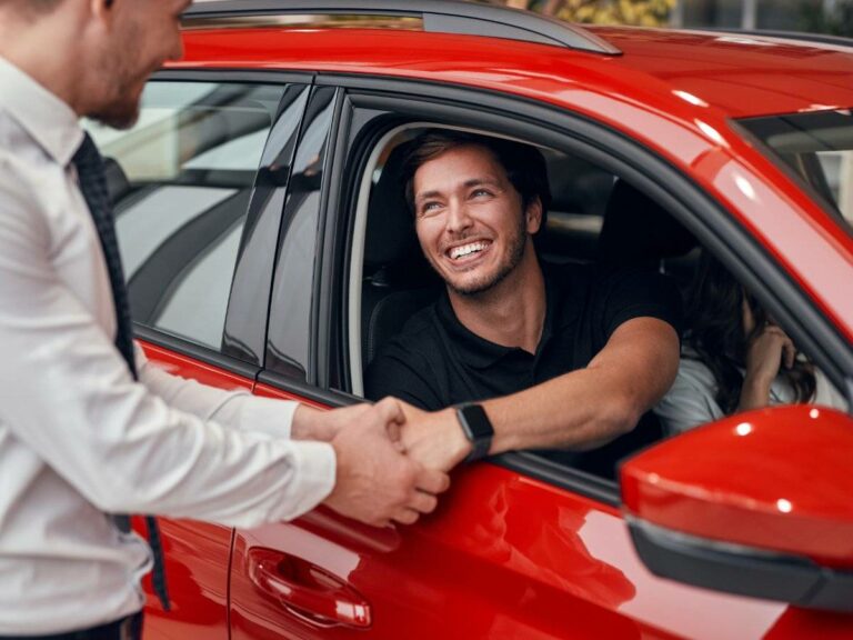 Happy customer sitting in a new red car, shaking hands with a car dealership representative.