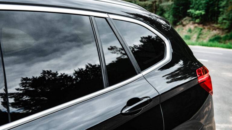 Close-up of the rear side window and door of a black SUV with tinted windows, reflecting trees and a cloudy sky.