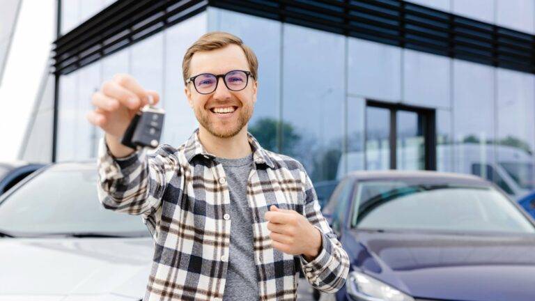 Smiling man holding car keys in front of a car dealership, standing near parked cars.