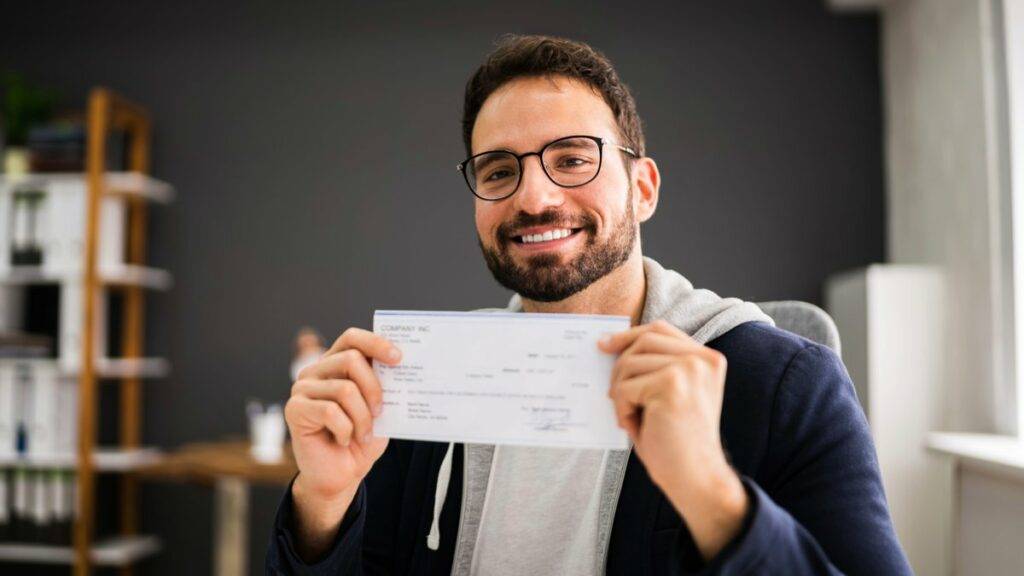 Man smiling while holding a company cheque in an office environment.