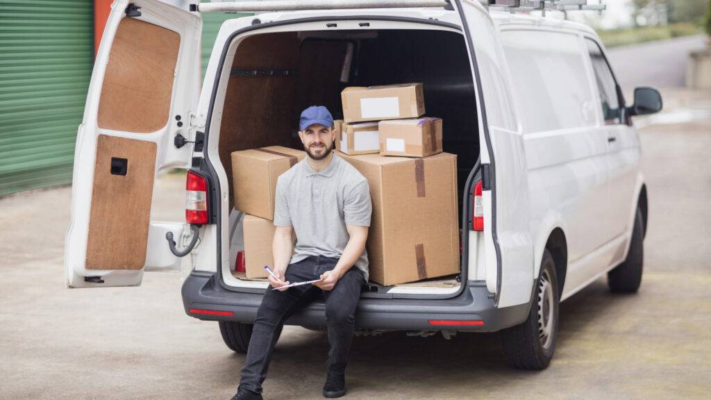 Delivery driver seated in a van's open cargo area with cardboard boxes and a clipboard.

